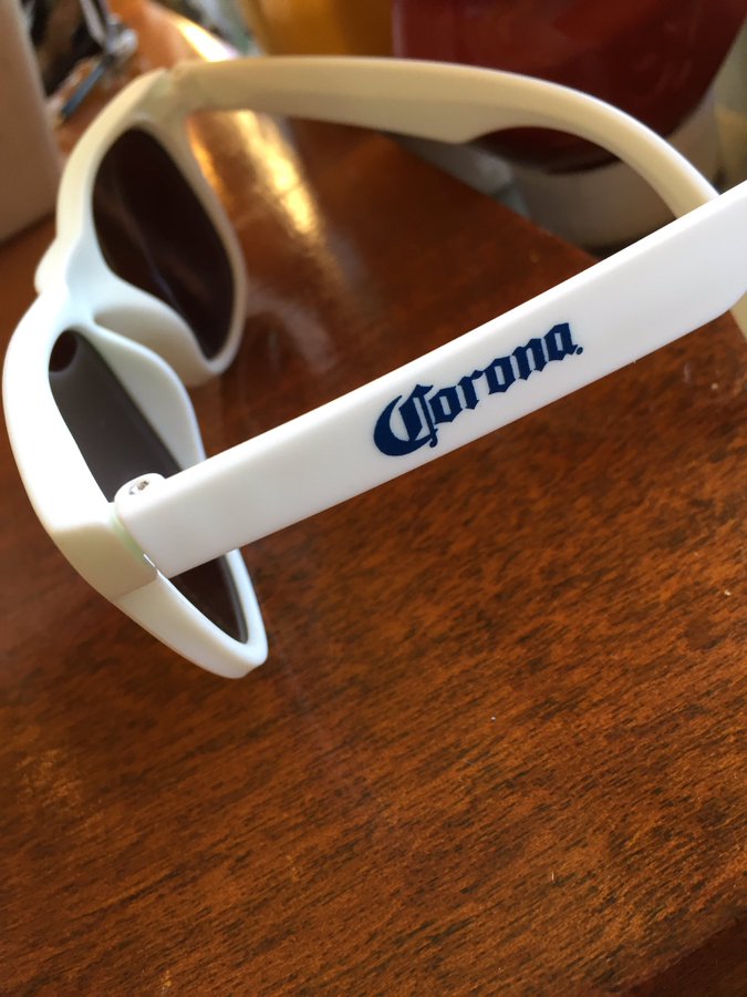 A pair of cheap, white sunglasses sitting on a desk. On one arm of the sunglasses, the blue logo of the Corona beer company is printed.