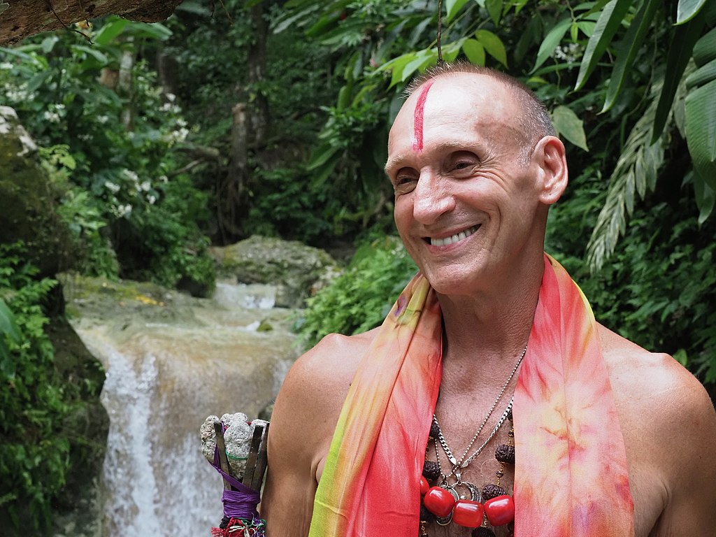 Shantaram's author standing in front of a waterfall with a jewelry necklace, a vertical line of red paint on his bald forehead, and a giant grin.