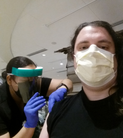 The author, masked and seated, receiving a vaccine from a syringe, which a nurse wearing a face shield holds with gloved hands and presses into the author's right arm.