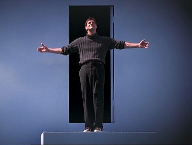 The image shows Jim Carrey as Truman in the movie. He's standing atop steps and before a door. He has his arms and hands spread wide, and he's looking up, smiling.