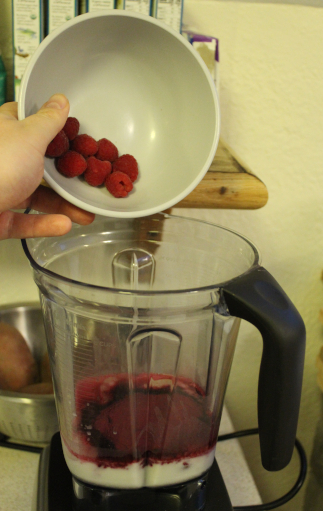 The photo shows the smoothie in progress. The photo shows my hand dumping a bowl of eight raspberries into the blender container.