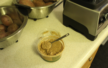 The photo shows, on a countertop, a tub of almond butter. In the background are the blender, red potatoes, and garnet yams.