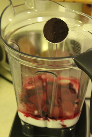 The photo shows the smoothie in progress. At the open top of the container, a tablespoon packed with maqui powder is poised to dump it in.