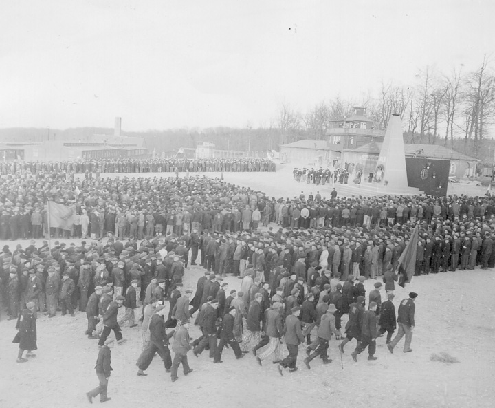 The black-and-white photograph shows the Buchenwald concentration camp in the bakground, with a rally in the foreground.