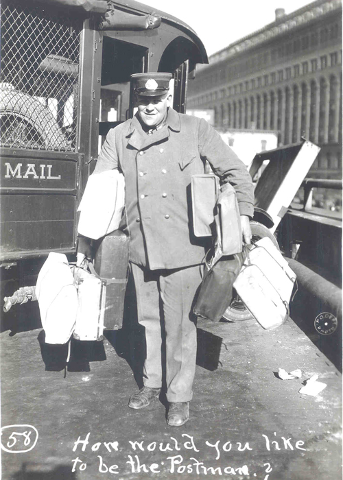 The black-and-white image shows a frontal view of a mailman. Both his hands are full carrying suitcases, and he has suitcases under his arms as well. He's smiling and he has a uniform hat on. In the background is his mail vehicle. Words at the bottom say in white, "How would you like to be the Postman?"