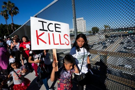 The image is a high quality photograph of protestors, primarily Hispanic, crossing a bridge above a freeway. One in front, a young boy, carries a stark sign that simply says "ICE Kills" in black and red against a white background.