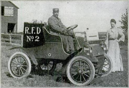 The black-and-white photo shows a uniformed man atop an old-timey automobile. A woman stands behind him. They're in some sort of rural farm setting.