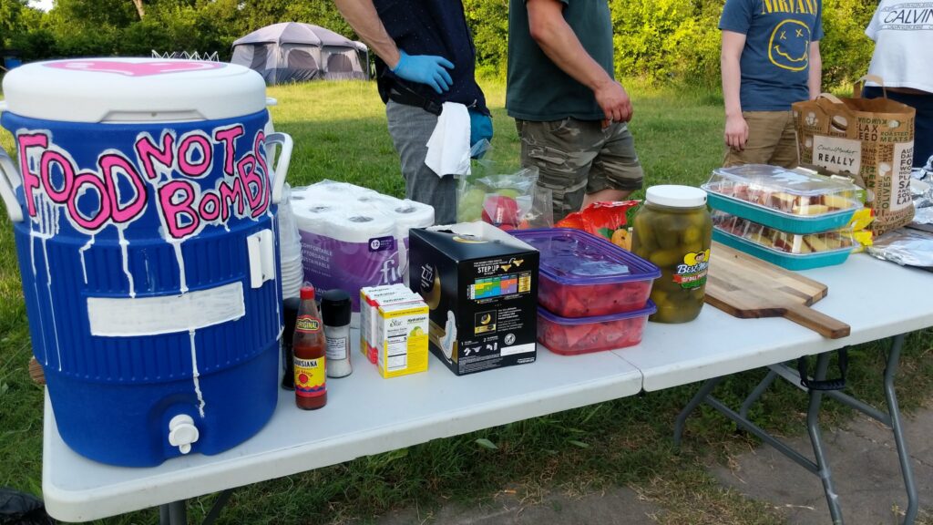 The image shows a folding table set up at a park. On the folding table is a blue water cooler with Food Not Bombs painted on it. Next to the cooler are various food items such as pickles and watermelon. In the background stand what I believe are four Food Not Bombs participants, one with camo pants, another with a Nirvana T-shirt.