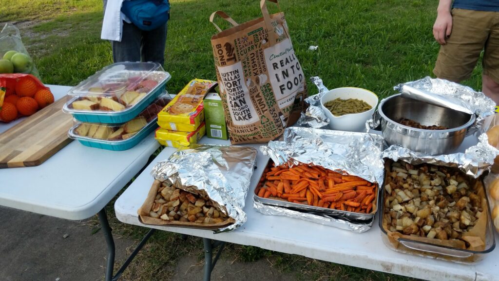 The image shows folding tables in a park with food items set atop. In the background stand what I believe are two members of Food Not Bombs Dallas.