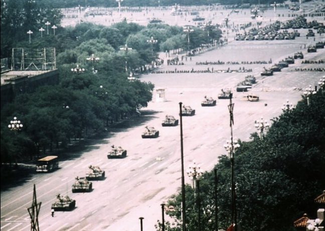 The image shows a man standing defiantly in front of a very long line of Chinese tanks