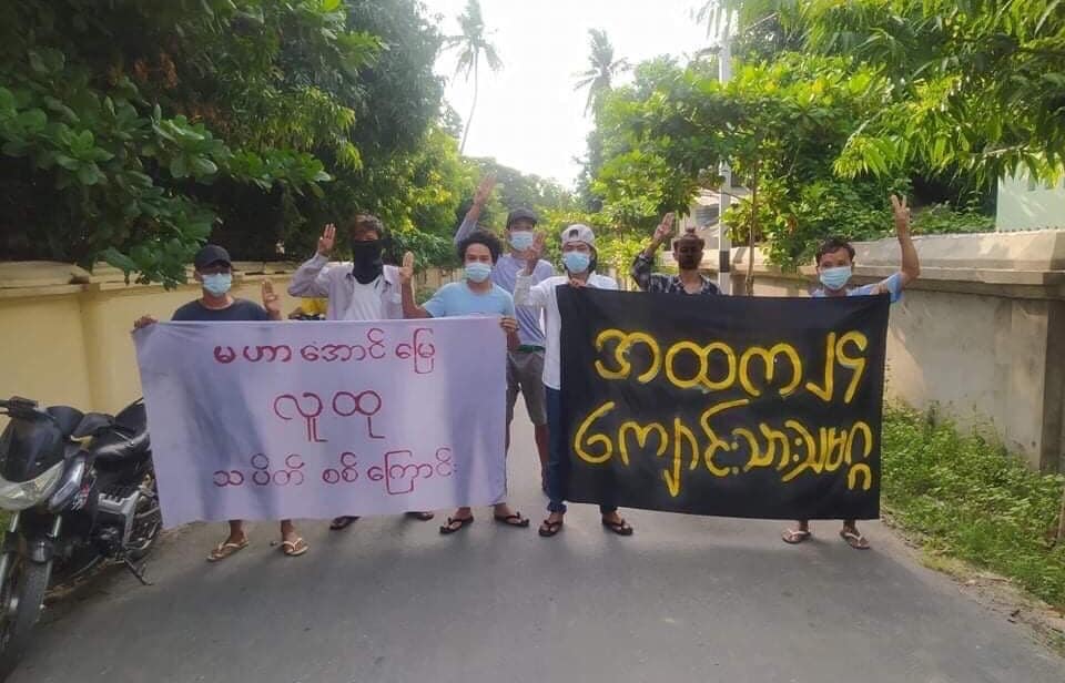 The image show a narrow roadway with a parked motorcycle, bordered by trees and walls. Standing on the roadway are young masked protesters making the three-finger democracy salute and holding signs with Burmese written on them.