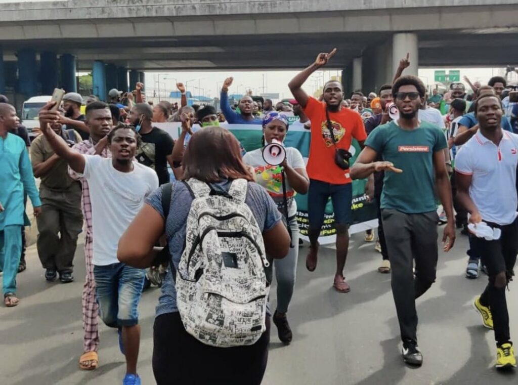 The colorful image shows protestors marching during the day in Nigeria, including one woman with a bullhorn, and many holding their hands in the air