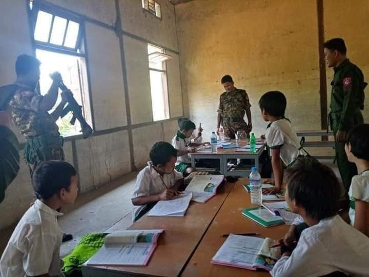 The photo shows a bare schoolroom. Elementary age children sit at tables unmasked, looking a workbooks or the soliders standing around, who are armed and taking the place of teachers