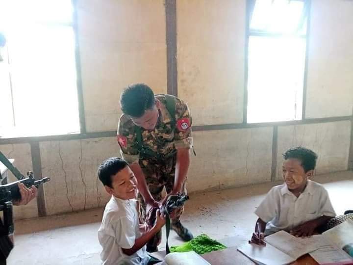 The photo, apparently from the same schoolroom as the above image, shows unmasked schoolkids at a desk in a bare classroom. A soldier, replacing a teacher, is showing one of the smiling schoolkids his gun.