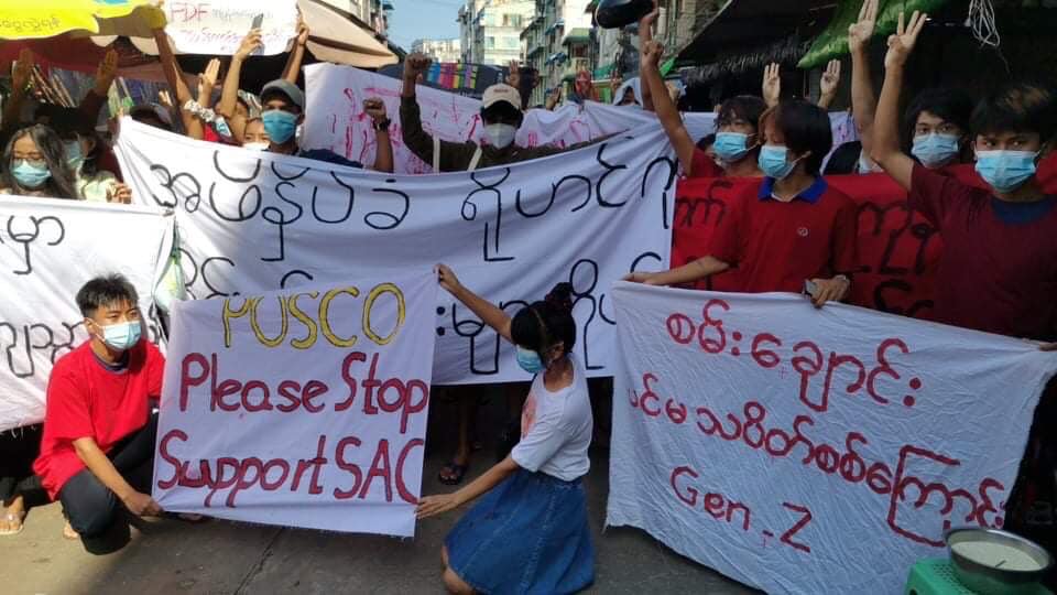 The image shows protesters in a market street of Yangon, some standing, some kneeling, all masked. They are holding signs, upon which is writing mostly in Burmese, else in English