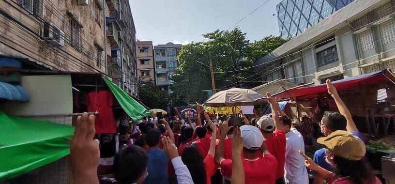 The image shows protestors marching through a market street in Yangon