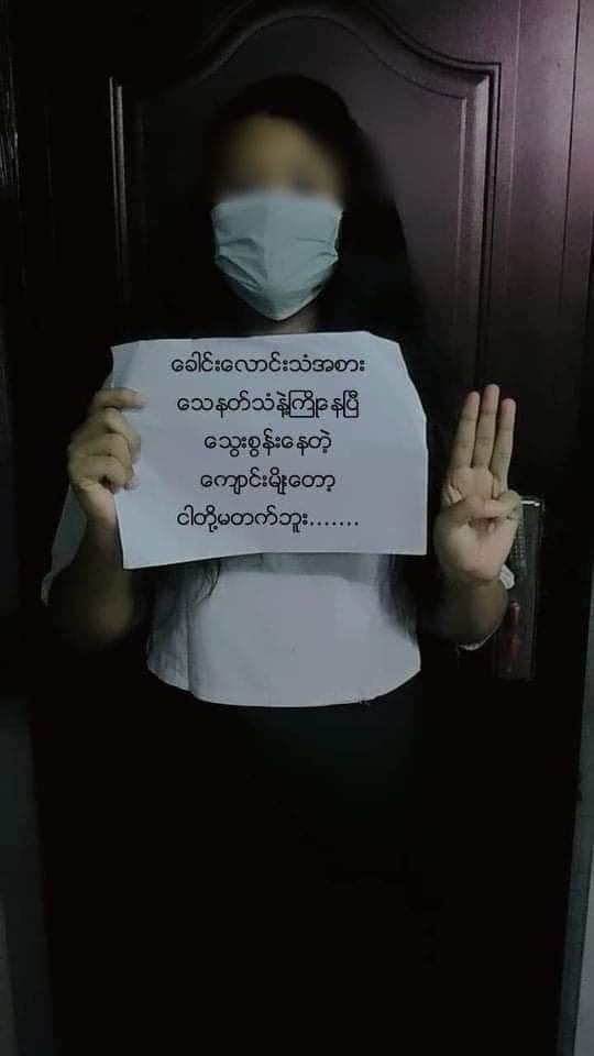 The image shows a masked young girl, perhaps early teens, with her face blurred. She's standing in front of a door and making the three-finger democracy salute. Her other hand holds a sign, upon which is writing in Burmese.