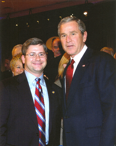 The color photo shows the two suits at some event or other. McHenry, wearing glasses, is smiling huge, probably showing his pleasure at being near one of the top dogs, George W. Bush. Bush II looks like it's just another day on the job of mugging for photos.