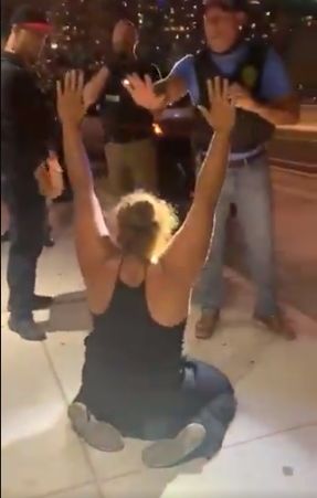 Piccini kneeling on the sidewalk, hands in the air, as the riot gear cops surround her and gesture at her.
