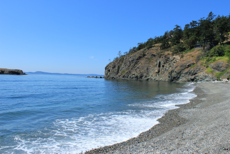 Idyllic color photo shows beach and ocean washing in below a clear blue sky. In the distance are hills, trees, etc.