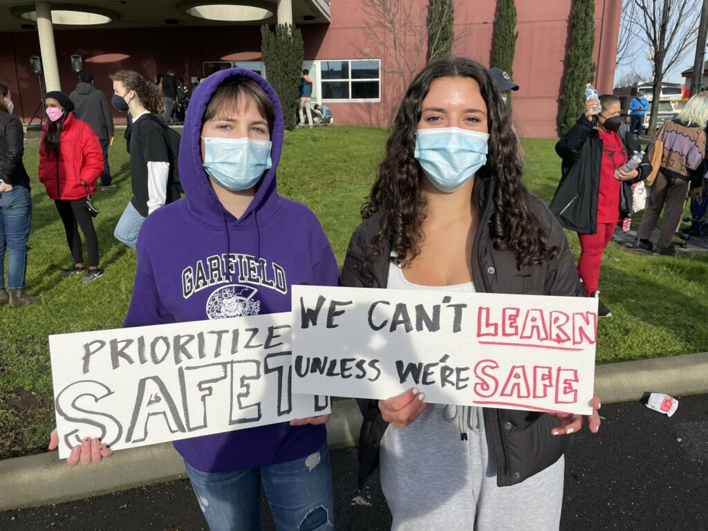 Two students holding protest signs. One reads: "Prioritize safety." The other says: "We can't learn unless we're safe."