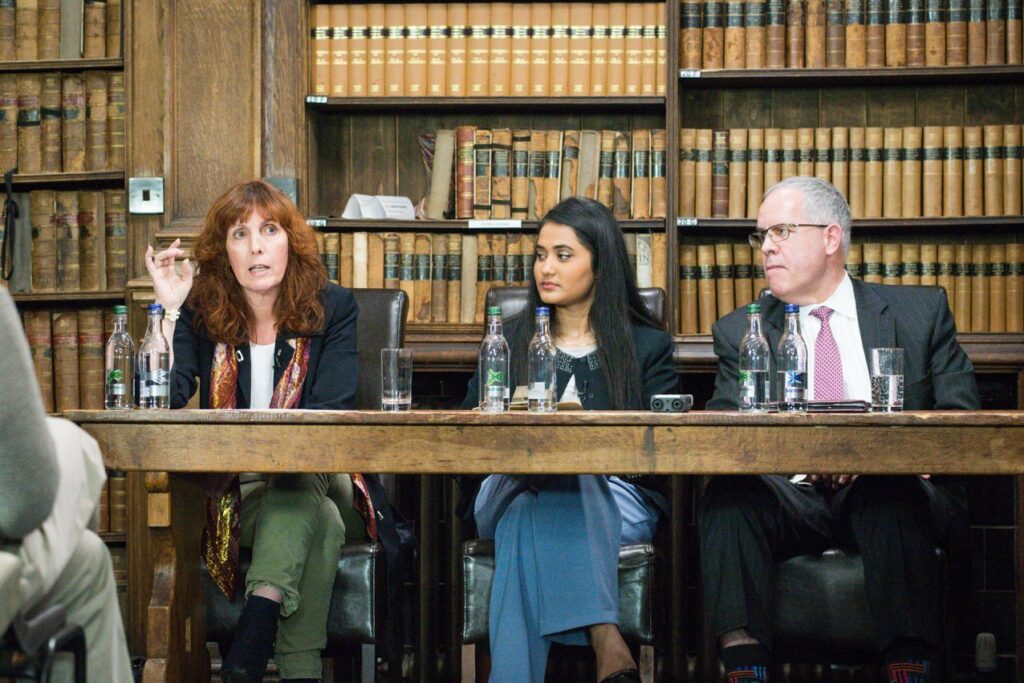 Shot of panel shows moderator watching Marsh with her hand raised, likely making a point, as Shedd looks on with angry expression.